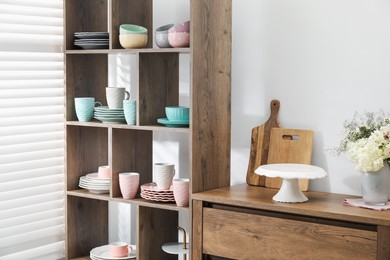 Photo of Shelving unit with different dishware, cups and floral decor near white wall indoors