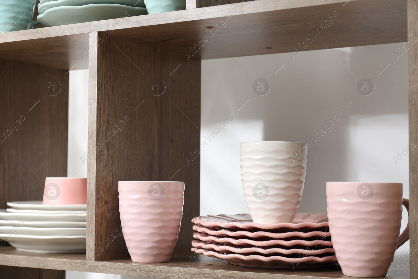Photo of Different plates and cups on shelves near white wall indoors, closeup