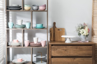 Photo of Shelving unit with different dishware, cups and floral decor near white wall indoors