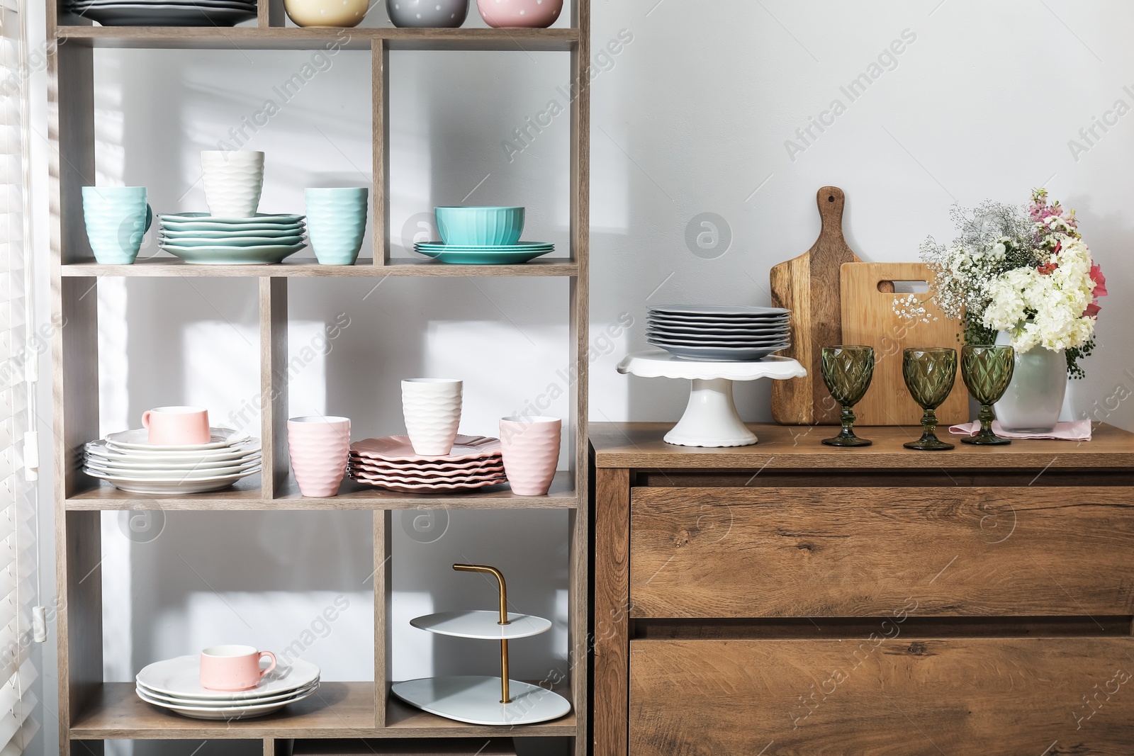 Photo of Shelving unit with different dishware, cups and floral decor near white wall indoors