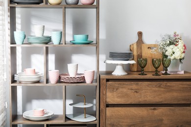 Photo of Shelving unit with different dishware, cups and floral decor near white wall indoors