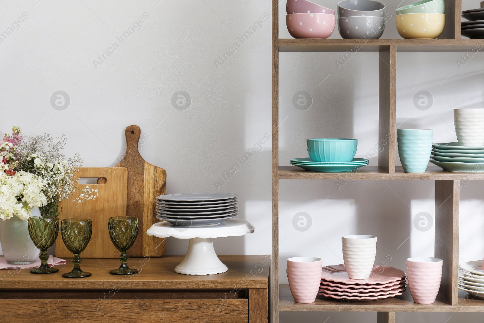 Photo of Shelving unit with different dishware, cups and floral decor near white wall indoors
