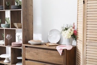 Photo of Shelving unit with different dishware, glasses and floral decor near white wall indoors