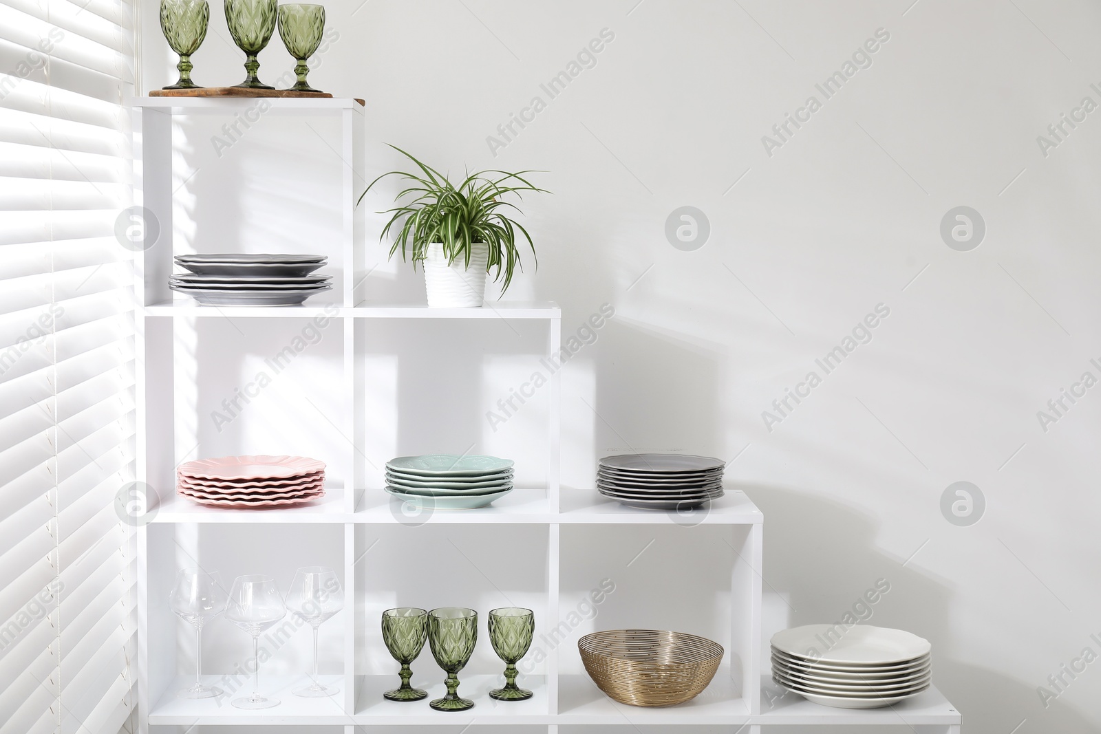 Photo of Shelving unit with different dishware, glasses and floral decor near white wall indoors