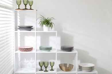 Photo of Shelving unit with different dishware, glasses and floral decor near white wall indoors