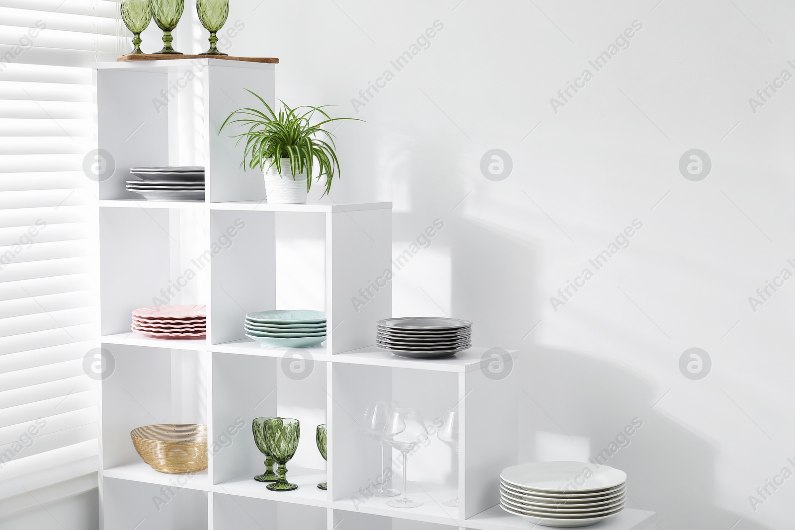 Photo of Shelving unit with different dishware, glasses and floral decor near white wall indoors