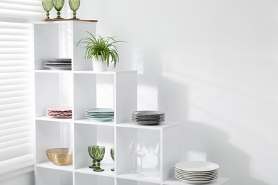 Photo of Shelving unit with different dishware, glasses and floral decor near white wall indoors
