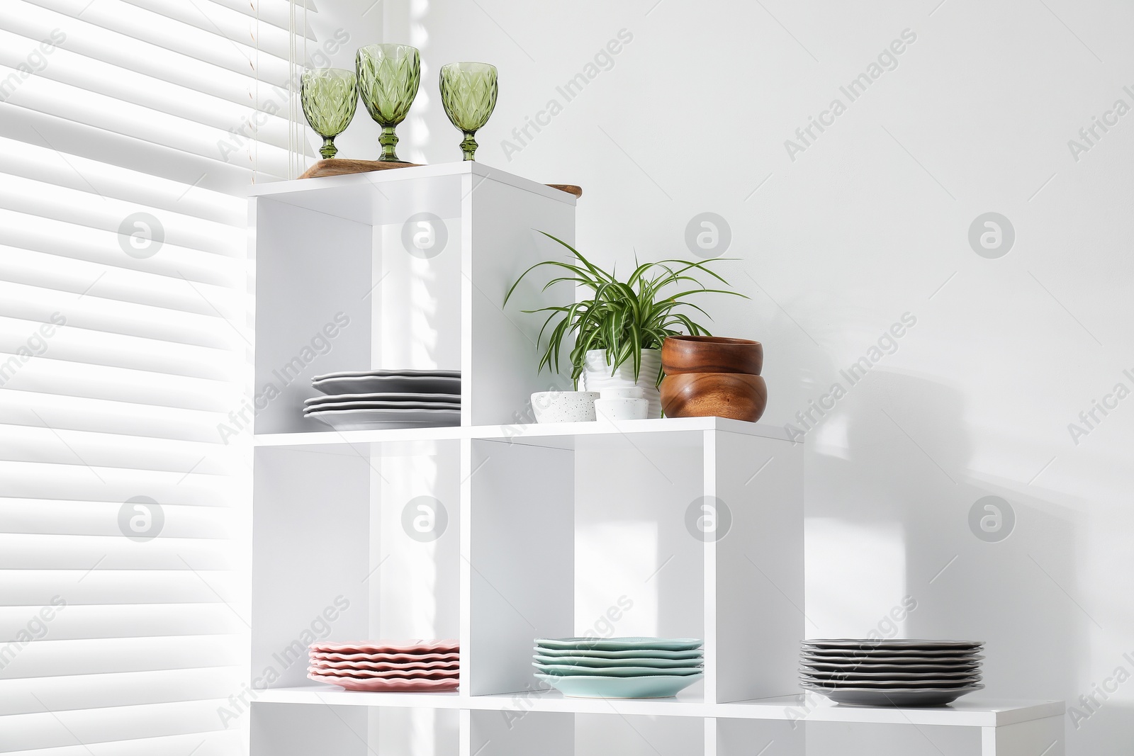 Photo of Shelving unit with different dishware, glasses and houseplant near white wall indoors, low angle view