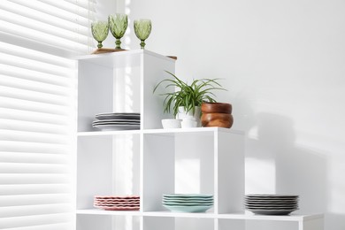Photo of Shelving unit with different dishware, glasses and houseplant near white wall indoors, low angle view