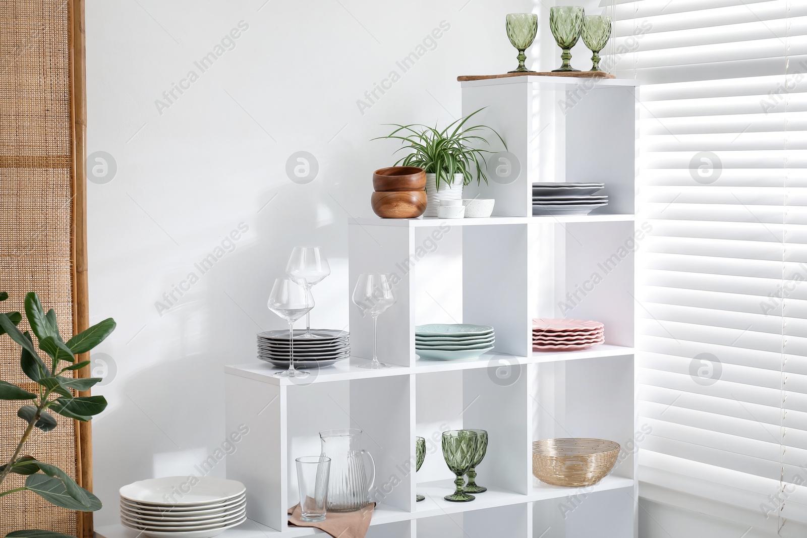 Photo of Shelving unit with different dishware, glasses and floral decor near white wall indoors