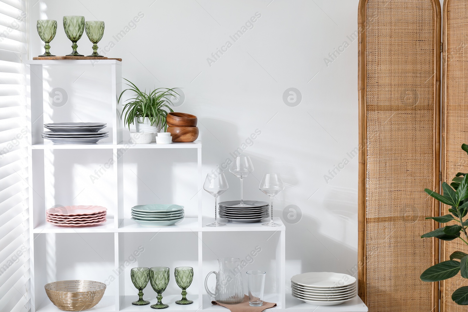Photo of Shelving unit with different dishware, glasses and floral decor near white wall indoors