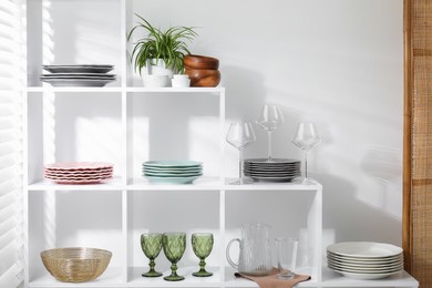 Photo of Shelving unit with different dishware, glasses and floral decor near white wall indoors