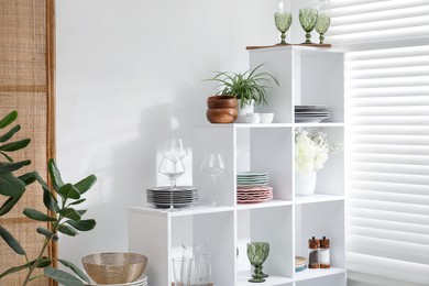 Photo of Shelving unit with different dishware, glasses and floral decor near white wall indoors