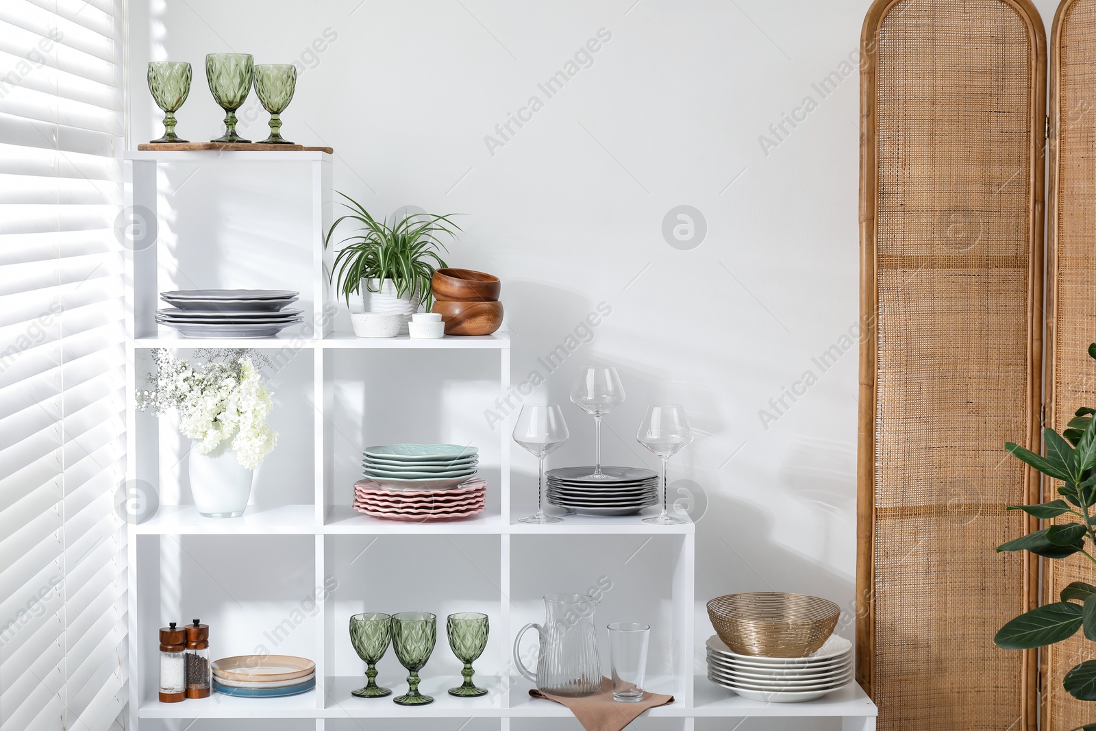 Photo of Shelving unit with different dishware, glasses and floral decor near white wall indoors