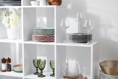 Photo of Shelving unit with different dishware, glasses and floral decor near white wall indoors