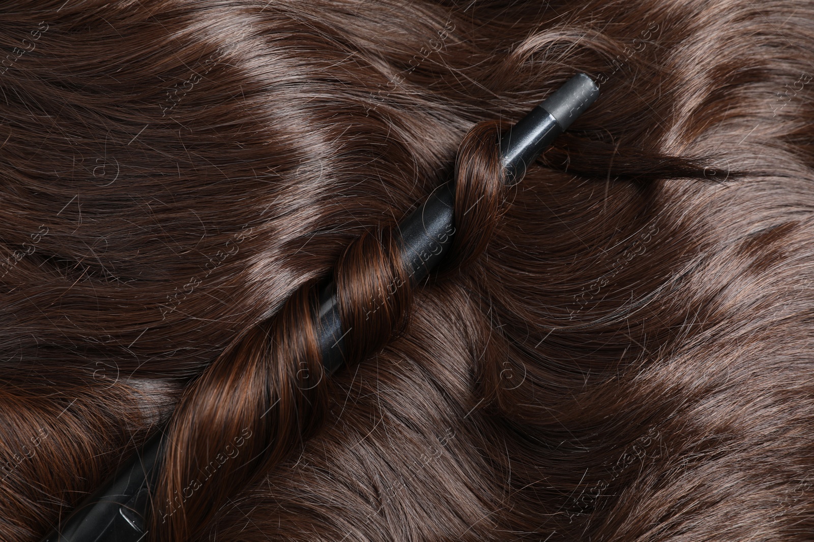 Photo of Curling iron with shiny brown hair, top view