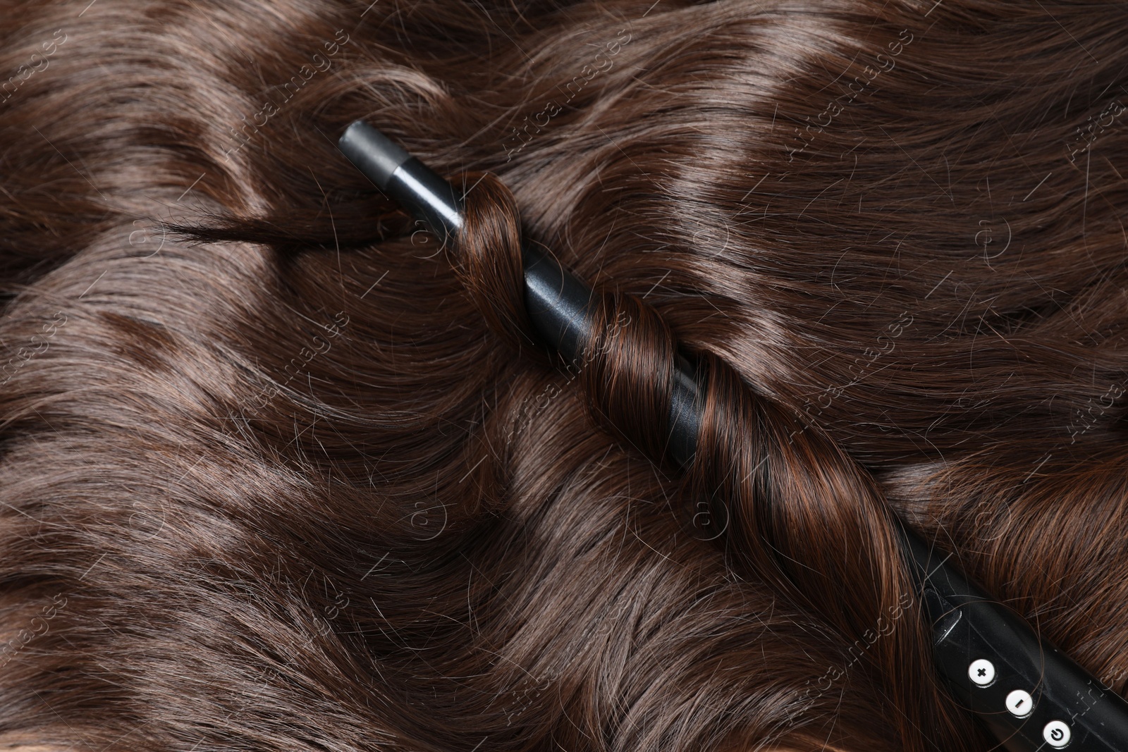 Photo of Curling iron with shiny brown hair, closeup