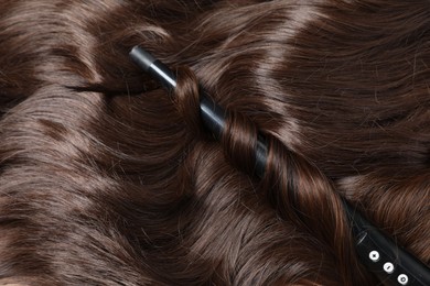 Photo of Curling iron with shiny brown hair, closeup