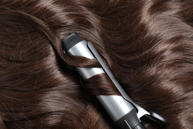 Photo of Curling iron with shiny brown hair, closeup