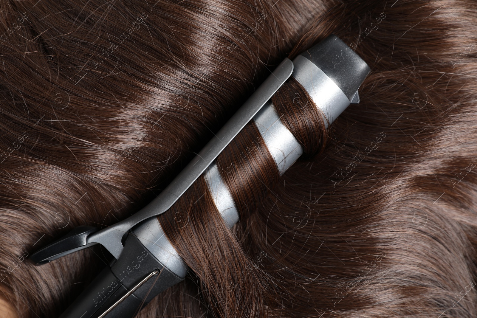 Photo of Curling iron with shiny brown hair, top view