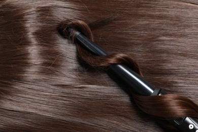 Photo of Curling iron with shiny brown hair, top view