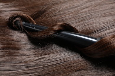Photo of Curling iron with shiny brown hair, closeup