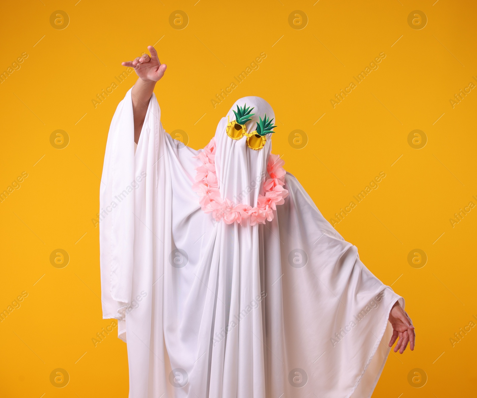 Photo of Funny ghost. Man covered with white sheet in sunglasses and wreath of flowers dancing on orange background