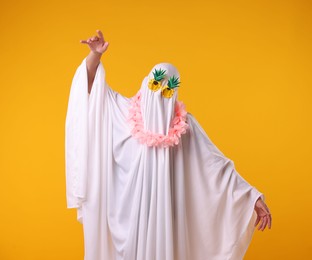 Photo of Funny ghost. Man covered with white sheet in sunglasses and wreath of flowers dancing on orange background