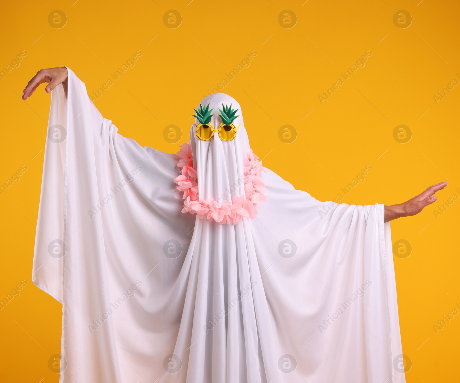 Photo of Funny ghost. Man covered with white sheet in sunglasses and wreath of flowers dancing on orange background
