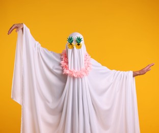 Photo of Funny ghost. Man covered with white sheet in sunglasses and wreath of flowers dancing on orange background
