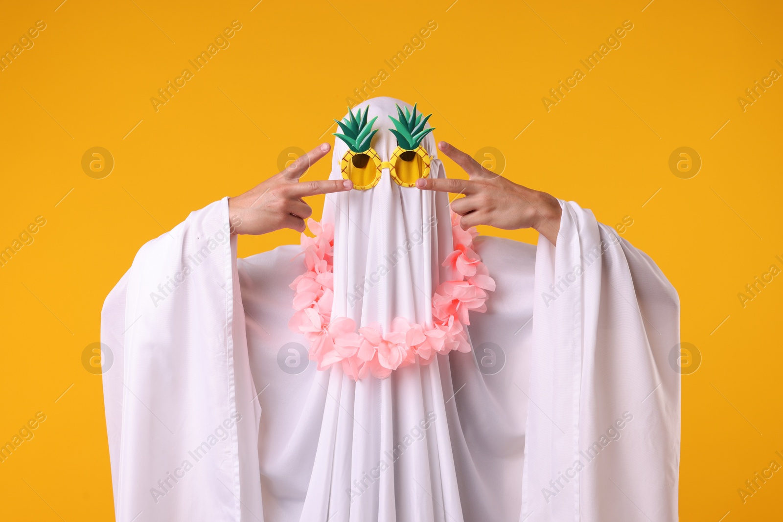 Photo of Funny ghost. Man covered with white sheet in sunglasses and wreath of flowers gesturing on orange background