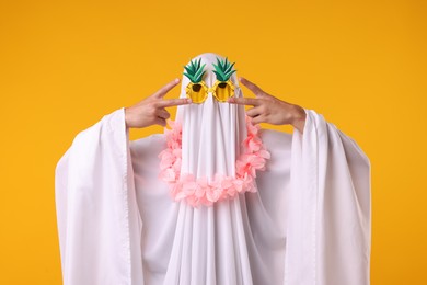 Photo of Funny ghost. Man covered with white sheet in sunglasses and wreath of flowers gesturing on orange background
