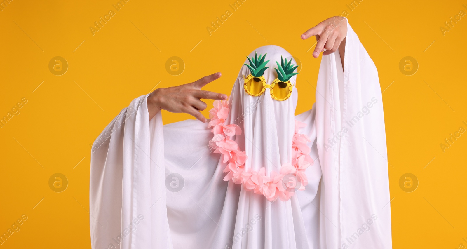 Photo of Funny ghost. Man covered with white sheet in sunglasses and wreath of flowers gesturing on orange background
