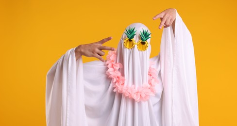 Photo of Funny ghost. Man covered with white sheet in sunglasses and wreath of flowers gesturing on orange background
