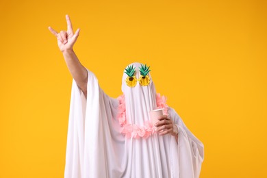 Photo of Man in ghost costume with sunglasses and wreath of flowers making rock gesture on orange background