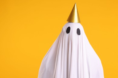 Photo of Funny ghost. Man covered with white sheet in party cone on orange background, space for text