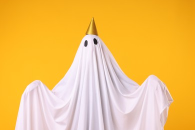 Photo of Funny ghost. Man covered with white sheet in party cone on orange background