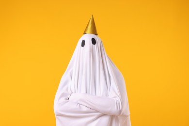 Photo of Funny ghost. Man covered with white sheet in party cone on orange background