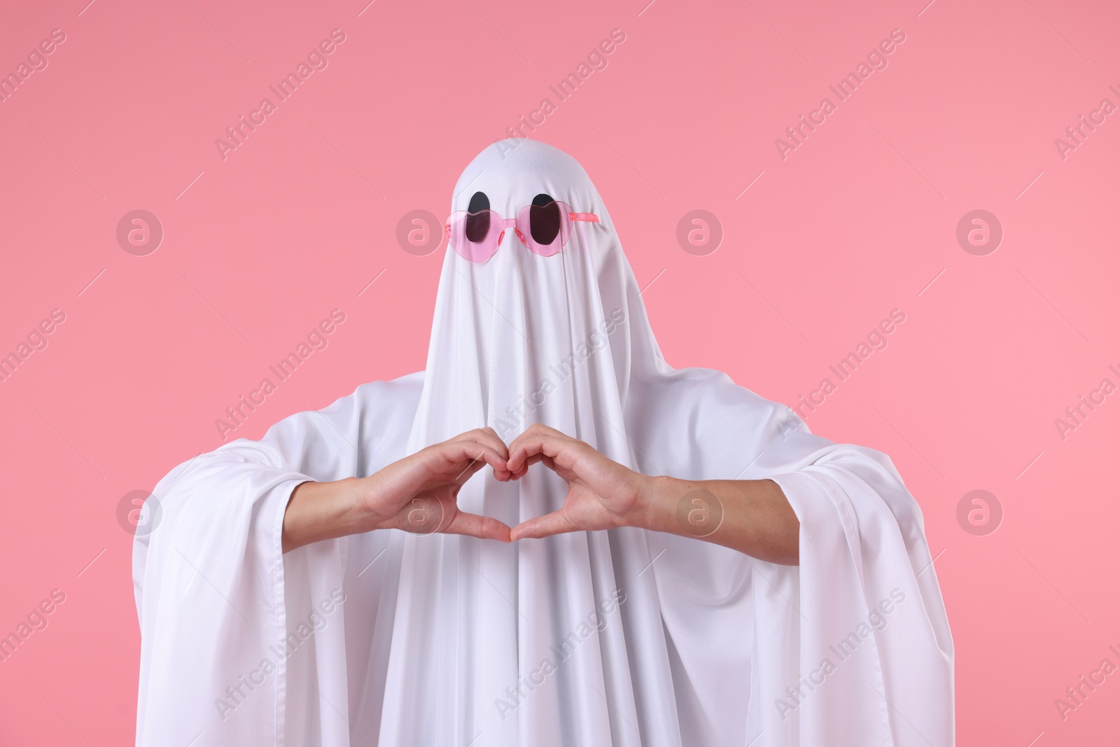 Photo of Cute ghost. Man covered with white sheet in sunglasses making heart gesture on pink background