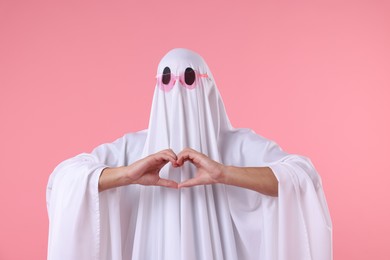 Photo of Cute ghost. Man covered with white sheet in sunglasses making heart gesture on pink background