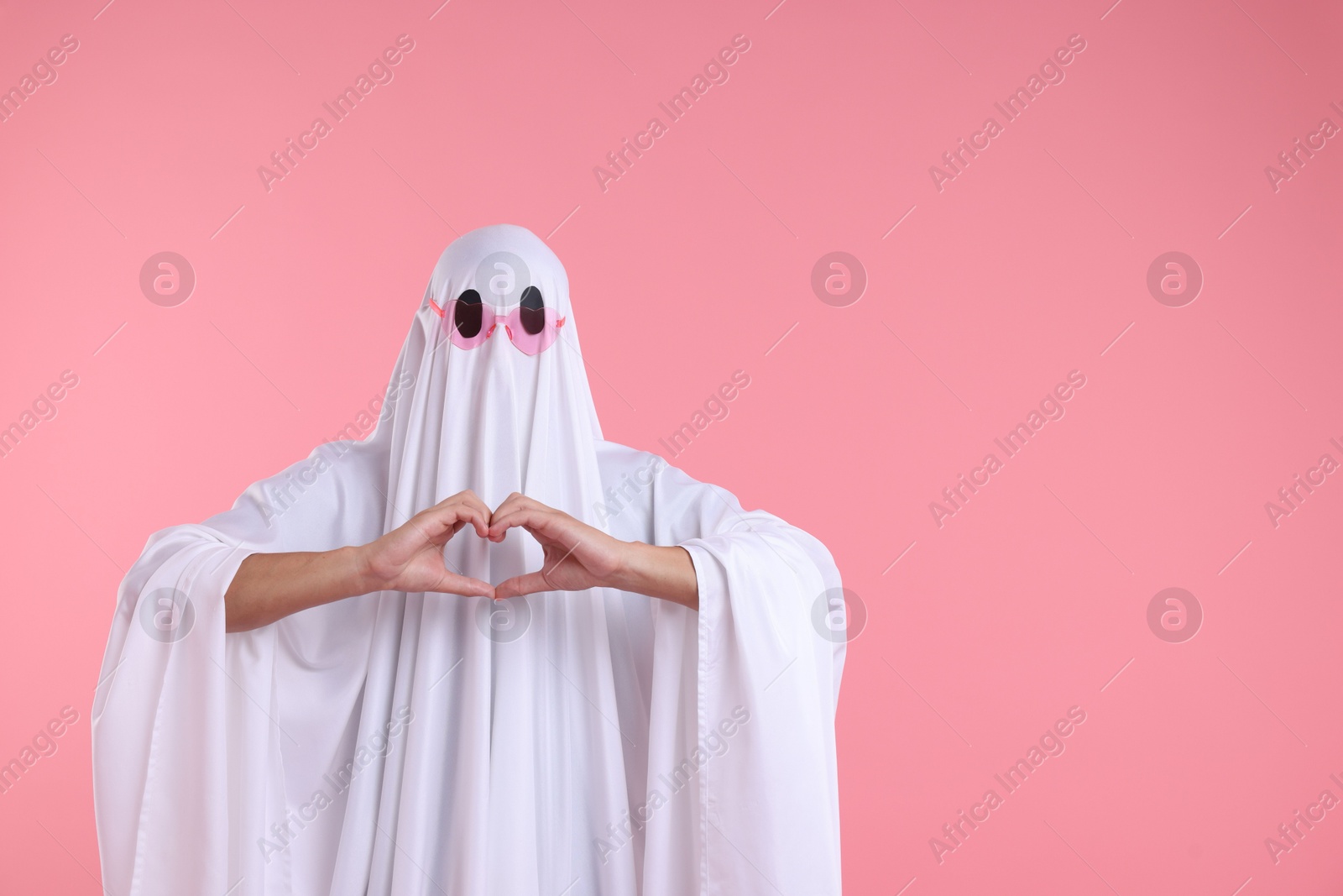 Photo of Cute ghost. Man covered with white sheet in sunglasses making heart gesture on pink background, space for text