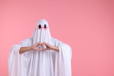 Photo of Cute ghost. Man covered with white sheet in sunglasses making heart gesture on pink background, space for text
