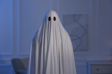 Photo of Creepy ghost. Man covered with white sheet in blue light at home