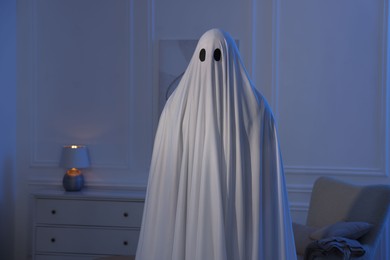 Photo of Creepy ghost. Man covered with white sheet in blue light at home
