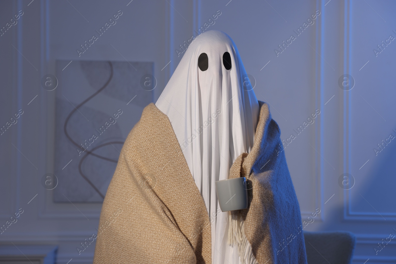 Photo of Man in ghost costume wrapped with blanket holding cup of drink at home