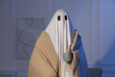 Photo of Man in ghost costume wrapped with blanket holding cup of drink at home