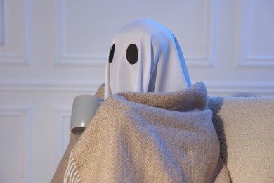 Photo of Man in ghost costume wrapped with blanket holding cup of drink at home