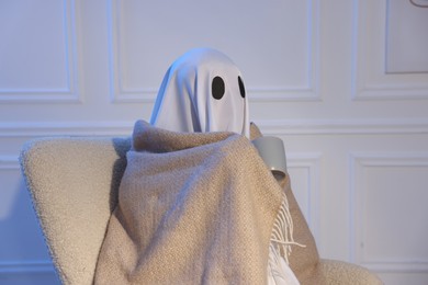 Photo of Man in ghost costume wrapped with blanket holding cup of drink at home