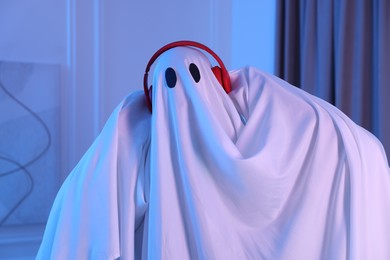 Photo of Funny ghost. Man covered with white sheet listening to music with headphones in blue light at home
