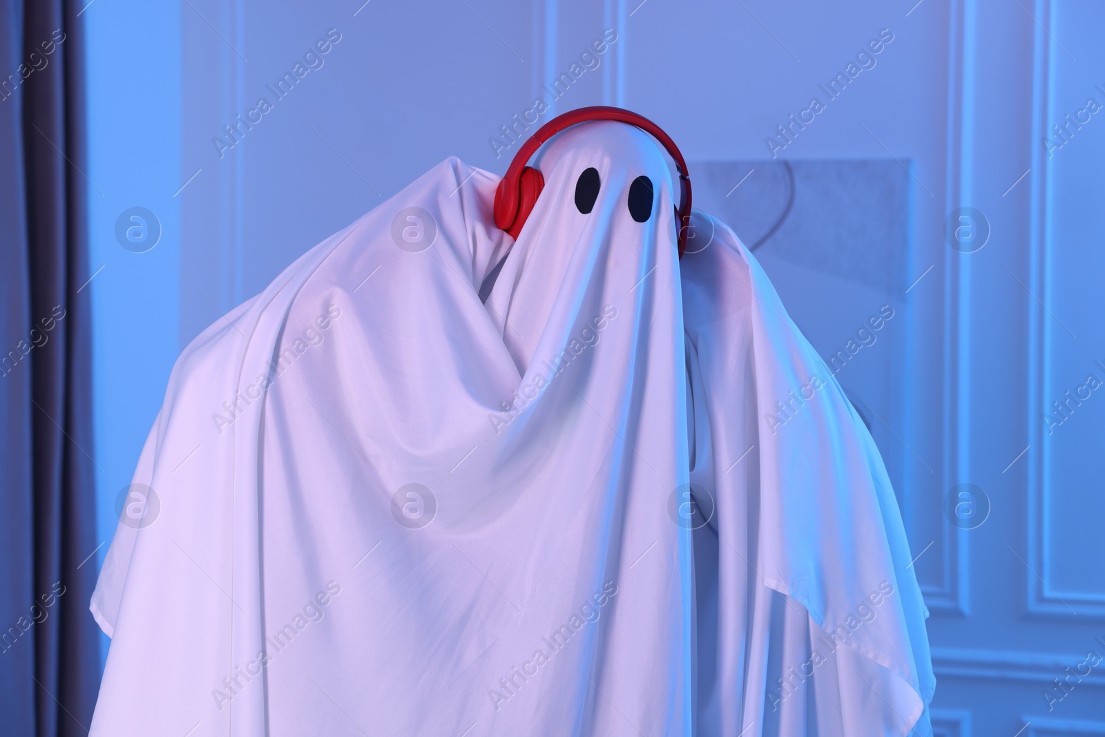 Funny ghost. Man covered with white sheet listening to music with headphones in blue light at home Photo of Funny ghost. Man covered with white sheet listening to music with headphones in blue light at home